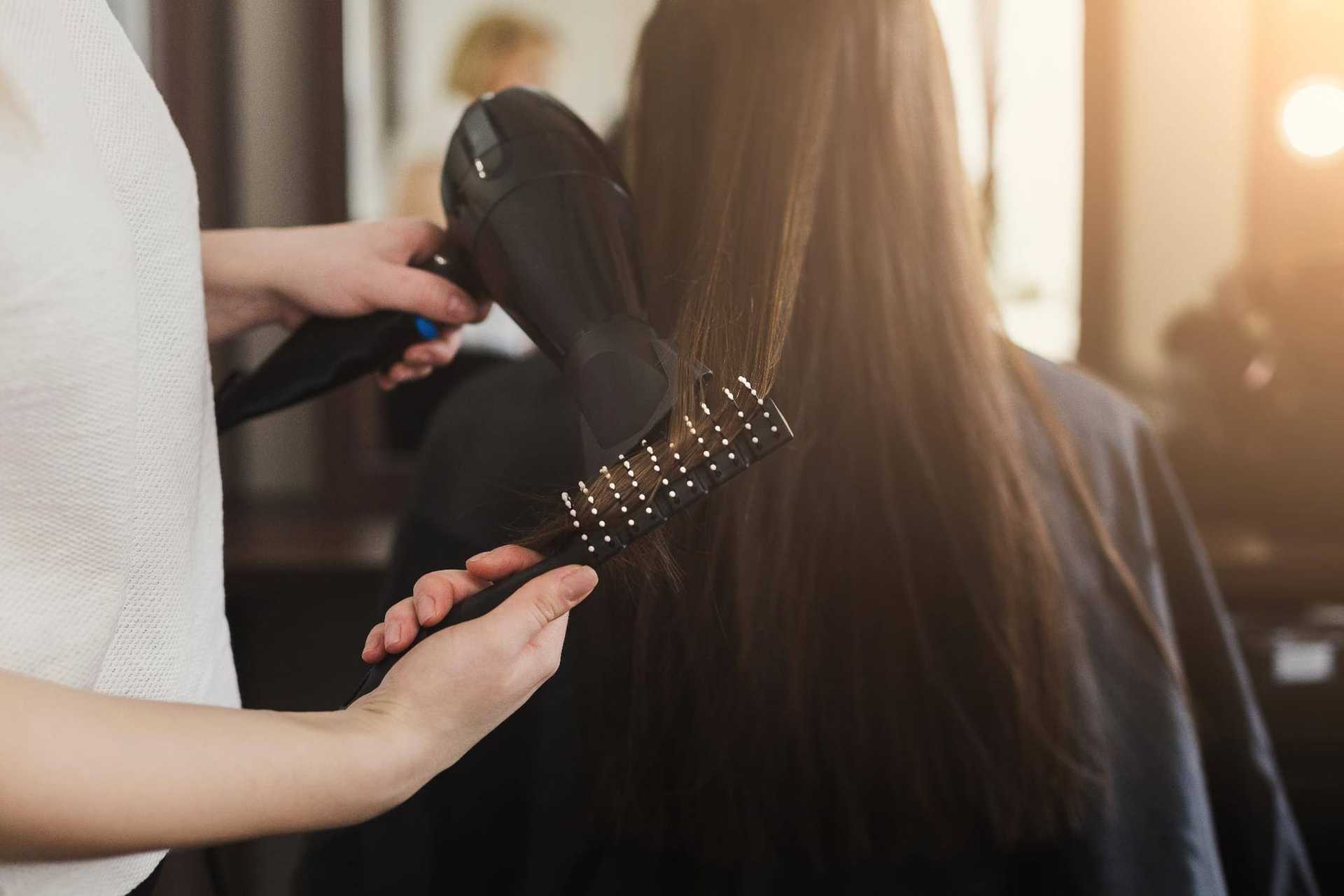 Hair stylist drying client's long brown hair in a salon using a brush and hairdryer.